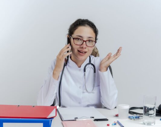 impressed young female doctor wearing medical robe and stethoscope and glasses sitting at desk with medical tools talking on phone looking at camera showing empty hand isolated on white background