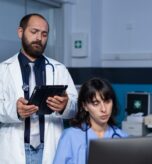 Team of medical workers using technology for overtime work. Doctor holding digital tablet for information while woman nurse looking at computer on desk for healthcare, working late.