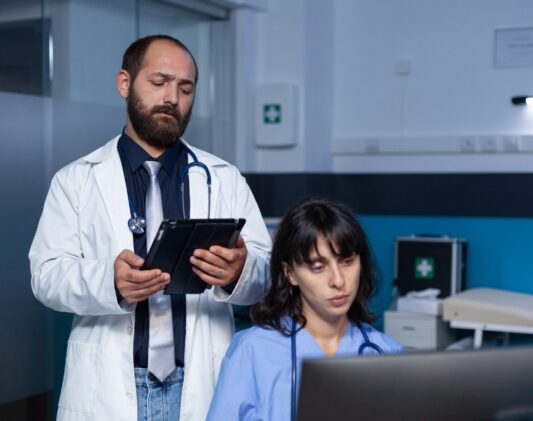 Team of medical workers using technology for overtime work Team of medical workers using technology for overtime work. Doctor holding digital tablet for information while woman nurse looking at computer on desk for healthcare, working late.