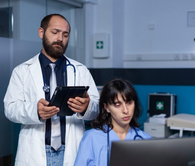 Team of medical workers using technology for overtime work. Doctor holding digital tablet for information while woman nurse looking at computer on desk for healthcare, working late.